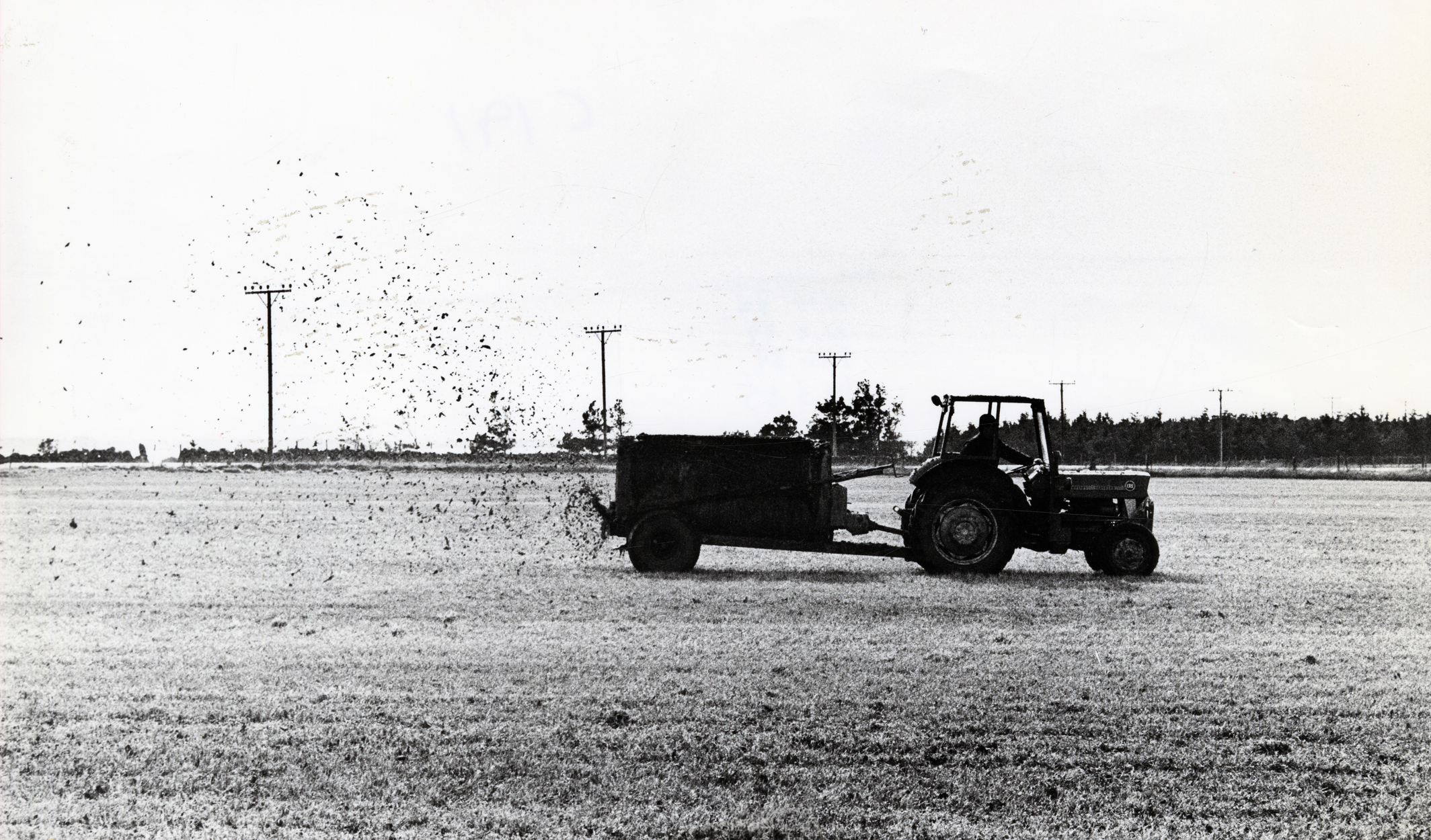 Et flatt jorde med en traktor med gjødselhenger. Både maskinene og gjødselen er i tydelig silhuett mot den lyse himmelen og marka.