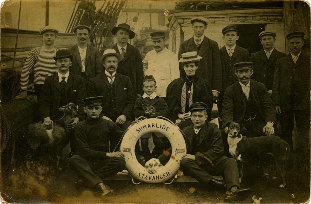 Mannskap bestående av 13 unge menn, en dame, en gutt, en hund og en geit (?) fotografert på dekk. Det står "Sumarlide - Stavanger" på livbøyen.