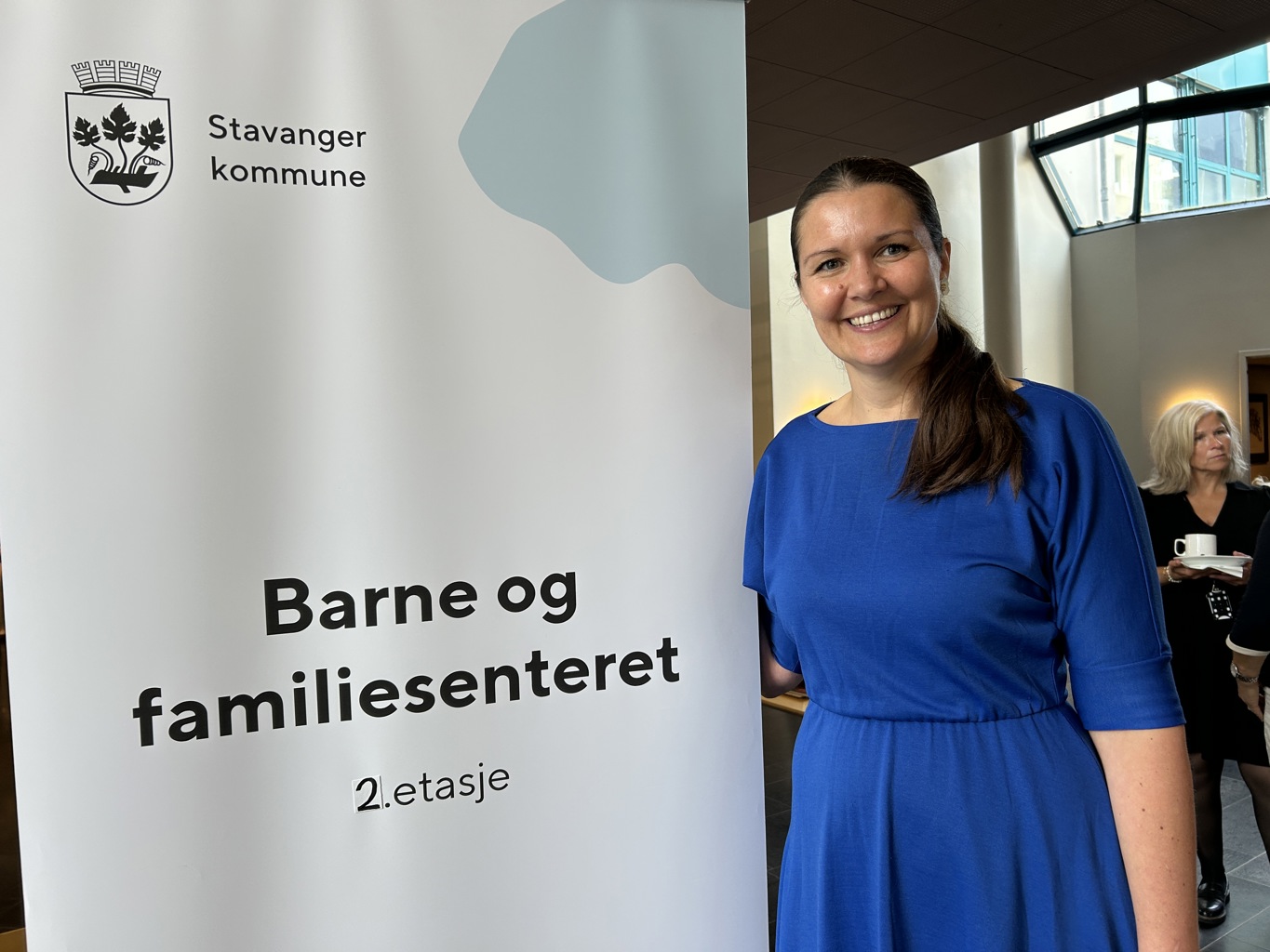 STOLT: En stolt virksomhetsleder Ingvild Rønneberg Hidle på åpningsdagen for barne- og familiesenteret. Ingvild Rønneberg Hidle står ved siden av et skilt med barne- og familiesenteret 2. etasje på. Hun har brukt hår, kongeblå kjole og et stort smil.